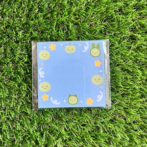 May include: A blue memo pad with a white border and a variety of green cartoon characters, including a cat, a bunny, and a melon. The characters are surrounded by yellow stars and white swirls.