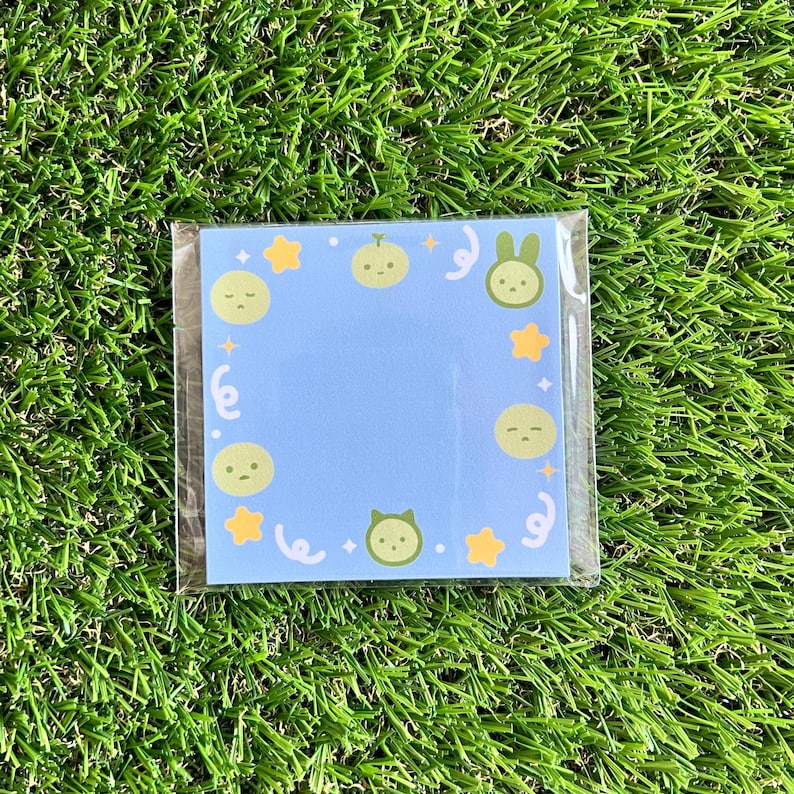 May include: A blue memo pad with a white border and a variety of green cartoon characters, including a cat, a bunny, and a melon. The characters are surrounded by yellow stars and white swirls.