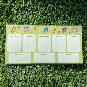 May include: A green weekly planner with pixelated food and star graphics. The days of the week are labeled, with space for notes. The planner is rectangular and sits on a bed of green grass.