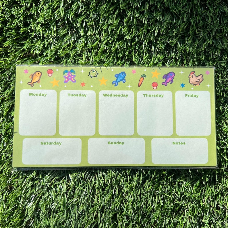 May include: A green weekly planner with pixelated food and star graphics. The days of the week are labeled, with space for notes. The planner is rectangular and sits on a bed of green grass.