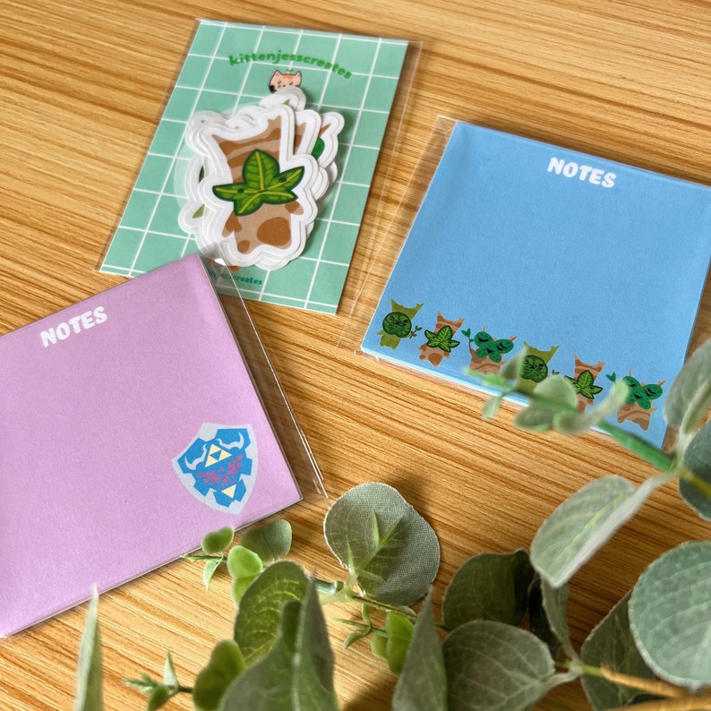 May include: Three sets of sticky notes with the word "NOTES" printed on them. The notes are in different colors: pink, blue, and white. The pink notes have a blue and pink sticker of a shield with a design on it. The blue notes have a row of green and brown stickers of a character with a leaf on its head. The white notes have a sticker of a brown character with a green leaf on its head.