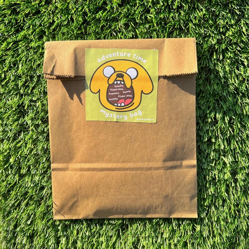 Op de afbeelding: Een bruine papieren zak met een gele sticker waarop een cartoonhond staat met de tekst "Adventure Time Mystery Bag" en een lijst met de meegeleverde artikelen: "Insides: Sticker pack, Bookmark, Buttons, Second sticker sheet".