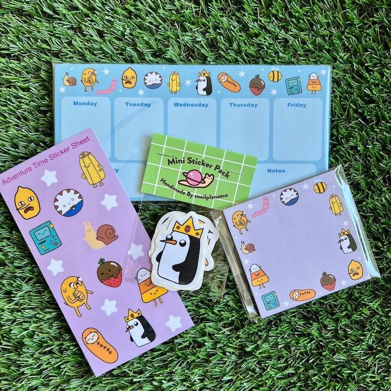 May include: A set of Adventure Time themed stickers featuring characters like Finn, Jake, and the Ice King. The set includes a sticker sheet, a mini sticker pack, and a notepad. The stickers are on a light purple background with white stars.