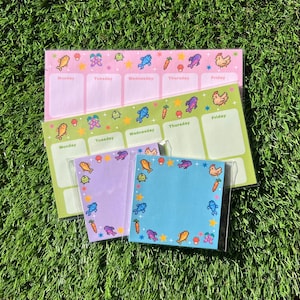May include: A collection of colorful weekly planner notepads with pixelated illustrations of fish, chickens, carrots, and mushrooms. The notepads are in shades of pink, green, purple, and blue, with days of the week labeled. The notepads are on a bed of green grass.