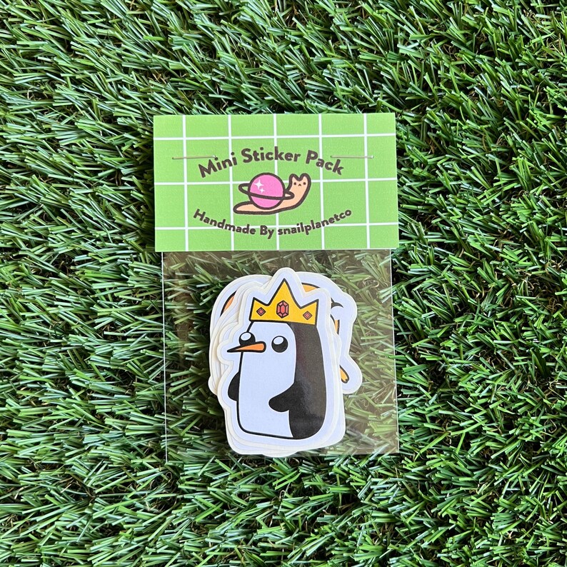 May include: A clear plastic bag containing a set of five mini stickers featuring a cartoon penguin wearing a crown. The stickers are white with black outlines and are packaged with a small card that reads "Mini Sticker Pack Handmade By snailplanetco".