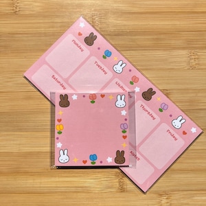 May include: Pink weekly planner and square notepad set. The planner has days of the week labeled and decorated with bunny faces, flowers, and stars. The notepad is framed with similar bunny and floral designs.