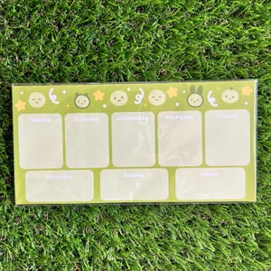 May include: A green weekly planner with a light green background and a white grid. The top of the planner has a row of cartoon characters with stars and moons. The days of the week are labeled: Monday, Tuesday, Wednesday, Thursday, Friday, Saturday, Sunday, and Notes.