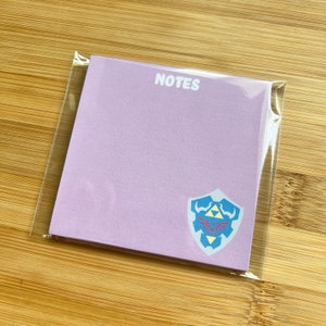 May include: A pack of lavender colored sticky notes with the word "NOTES" printed in white at the top. The notes feature a blue and yellow Triforce symbol on a white shield.