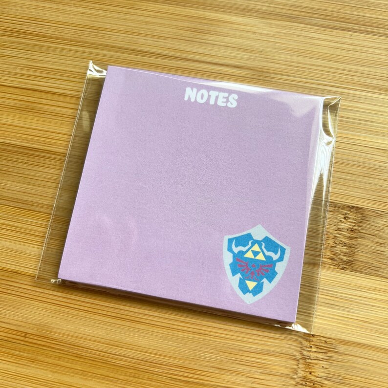 May include: A pack of lavender colored sticky notes with the word "NOTES" printed in white at the top. The notes feature a blue and yellow Triforce symbol on a white shield.