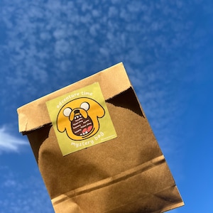 Op de afbeelding: Een bruine papieren zak met een gele sticker waarop een cartoonhond staat met de tekst "Adventure Time Mystery Bag" en een lijst met de items die erin zitten.