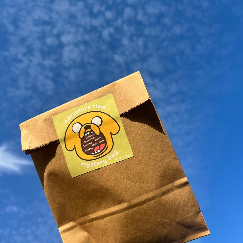 Op de afbeelding: Een bruine papieren zak met een gele sticker waarop een cartoonhond staat met de tekst "Adventure Time Mystery Bag" en een lijst met de items die erin zitten.