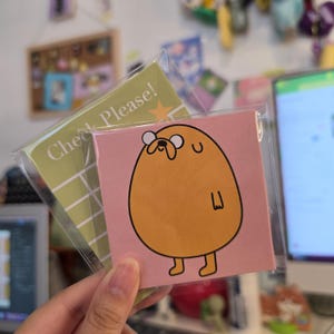May include: A square pink sticky note featuring a cartoon illustration of a round orange dog character with white eyes and a black outline. Behind it is a green rectangular notepad with white grid lines and the text Check Please! in white.