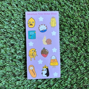 May include: A sticker sheet featuring characters from the animated television series Adventure Time. The sheet includes Finn the Human, Jake the Dog, Princess Bubblegum, Marceline the Vampire Queen, BMO, and more. The stickers are printed on a pink background with white stars.