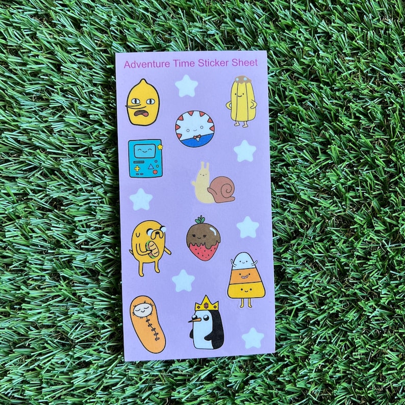 May include: A sticker sheet featuring characters from the animated television series Adventure Time. The sheet includes Finn the Human, Jake the Dog, Princess Bubblegum, Marceline the Vampire Queen, BMO, and more. The stickers are printed on a pink background with white stars.