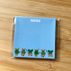 May include: A blue notepad with the word "NOTES" printed in white at the top. The bottom edge of the notepad features a row of seven cartoon characters with green leaves and brown bodies.