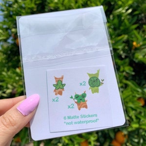 May include: A clear plastic bag containing six matte stickers featuring cartoon illustrations of green and brown creatures with leaves. The stickers are not waterproof and are labeled "6 Matte Stickers *not waterproof*"