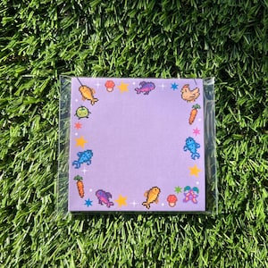 May include: A square, pixelated notepad with a light purple background. The notepad is decorated with colorful pixel art of fish, carrots, mushrooms, stars, and other whimsical elements. The notepad is encased in a clear plastic sleeve.