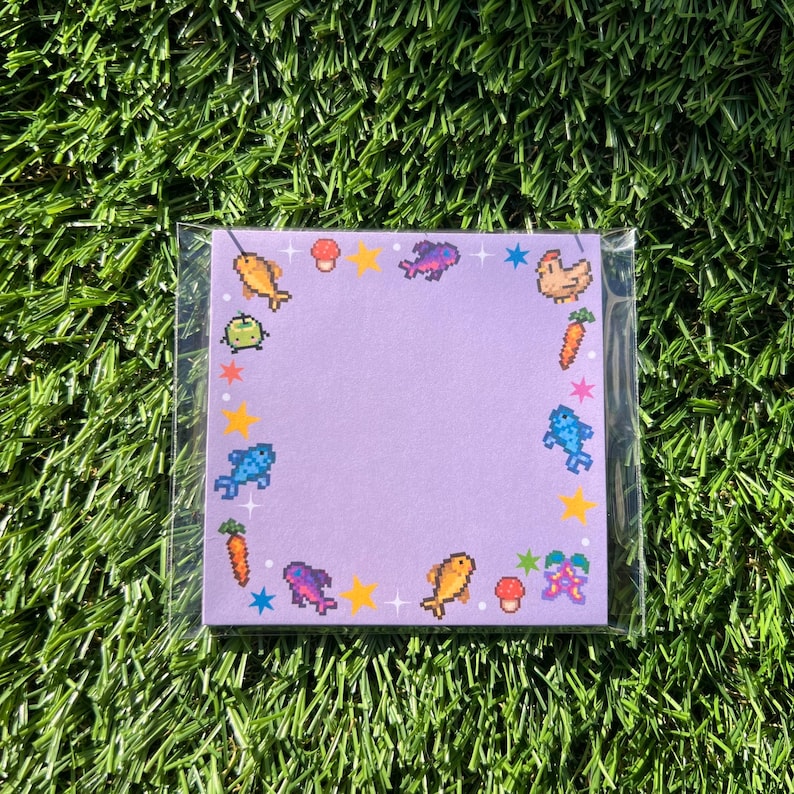 May include: A square, pixelated notepad with a light purple background. The notepad is decorated with colorful pixel art of fish, carrots, mushrooms, stars, and other whimsical elements. The notepad is encased in a clear plastic sleeve.