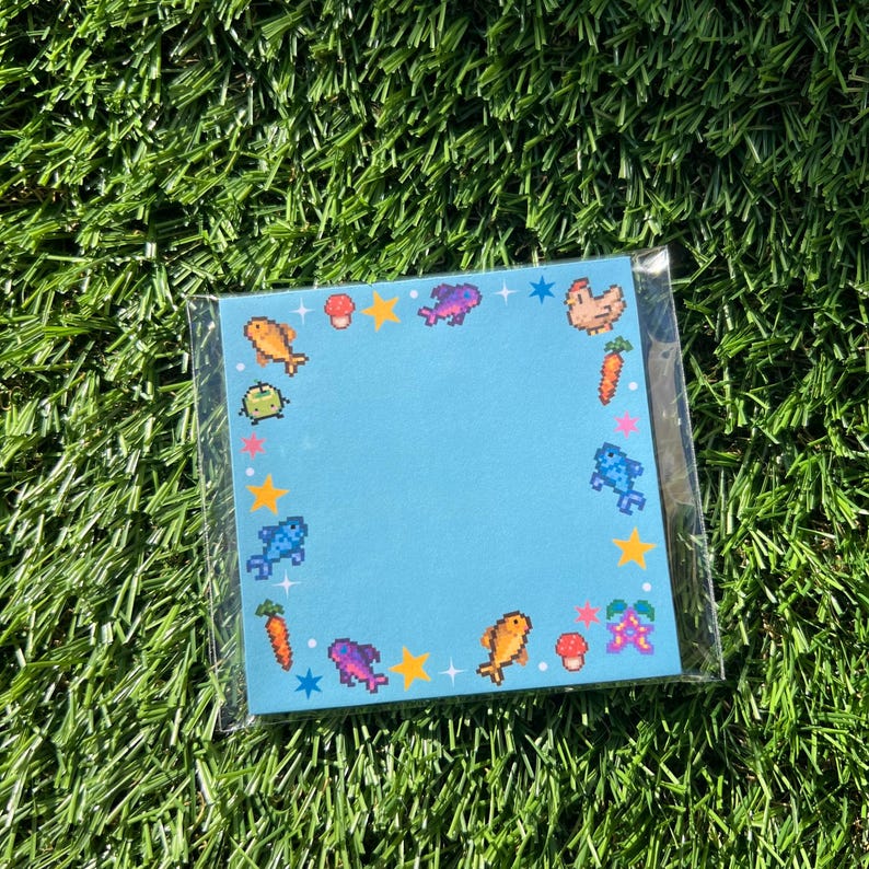 May include: A square, light blue notepad with pixelated illustrations of fish, mushrooms, stars, and other objects. The notepad is in a clear plastic sleeve, resting on a bed of green grass.
