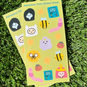 May include: A green sticker sheet with the text "Adventure Time Sticker Sheet" at the top. The sheet features various characters and objects from the Adventure Time cartoon, including Finn, Jake, Princess Bubblegum, BMO, and the Ice King. The stickers are colorful and have a cartoon style.