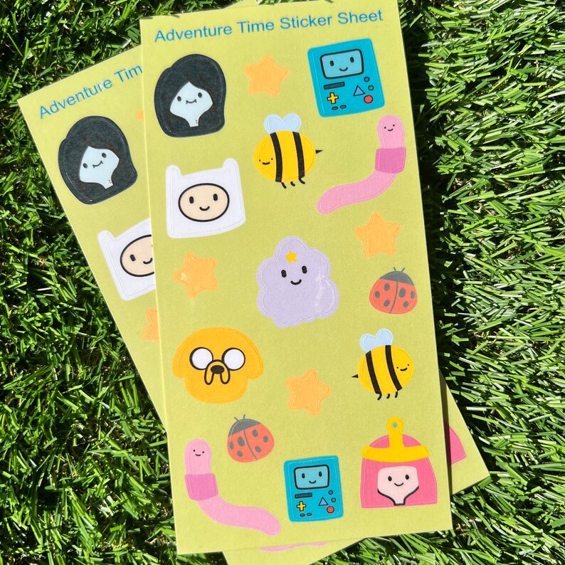 May include: A green sticker sheet with the text "Adventure Time Sticker Sheet" at the top. The sheet features various characters and objects from the Adventure Time cartoon, including Finn, Jake, Princess Bubblegum, BMO, and the Ice King. The stickers are colorful and have a cartoon style.