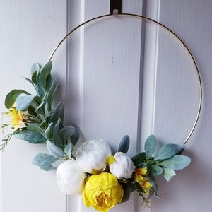 May include: A gold metal wreath with white and yellow flowers and green leaves. The wreath is hanging on a white door.