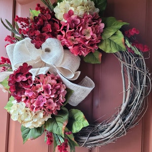 Puede incluir: Una corona de vid decorada con hortensias rosas y blancas, cinta de arpillera y ramitas de bayas rojas.