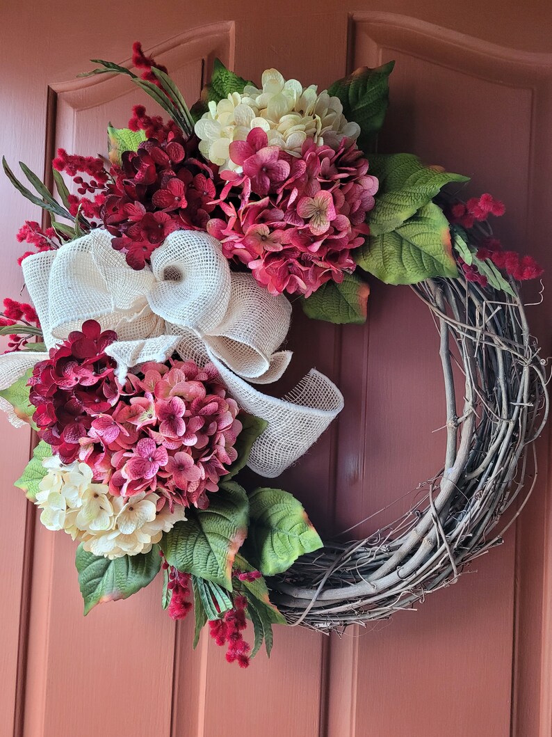Puede incluir: Una corona de vid decorada con hortensias rosas y blancas, cinta de arpillera y ramitas de bayas rojas.