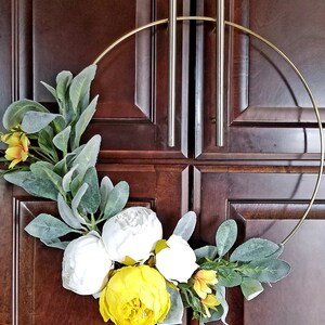 May include: A gold metal wreath with white and yellow artificial flowers and green leaves. The wreath is hanging on a wooden door.
