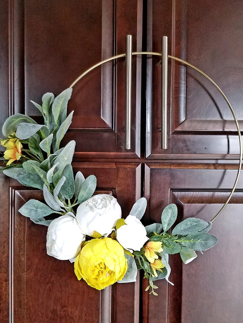 May include: A gold metal wreath with white and yellow artificial flowers and green leaves. The wreath is hanging on a wooden door.