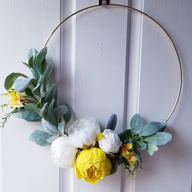 May include: A gold metal hoop wreath decorated with white and yellow artificial flowers and green leaves.