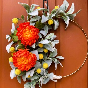 May include: A gold metal wreath with orange and yellow flowers and gray-green leaves. The wreath is hanging on a brown door.