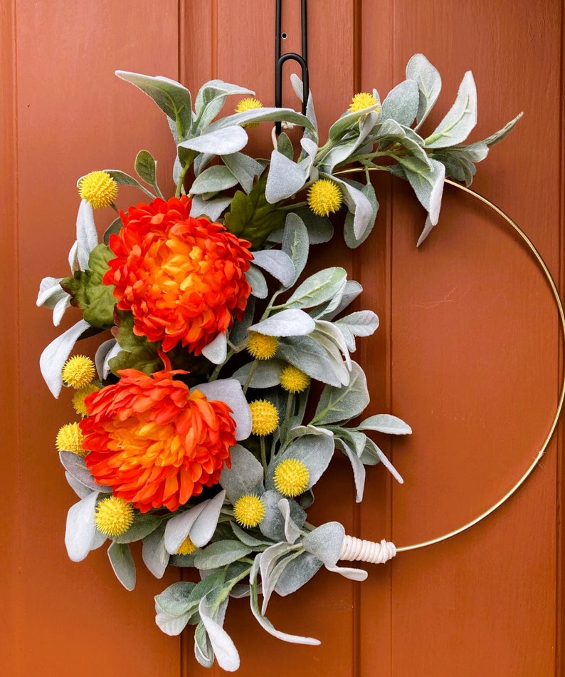 May include: A gold metal wreath with orange and yellow flowers and gray-green leaves. The wreath is hanging on a brown door.