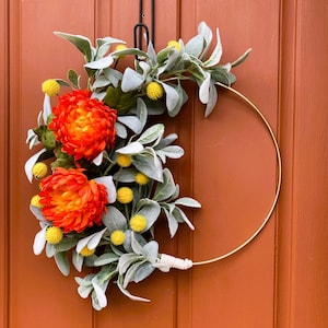 May include: A gold metal wreath with a white twine bow. The wreath is decorated with orange and yellow flowers and gray-green leaves.