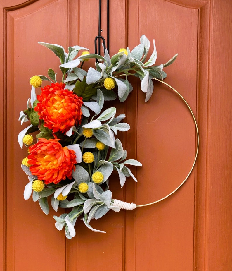 May include: A gold metal wreath with a white twine bow. The wreath is decorated with orange and yellow flowers and gray-green leaves.