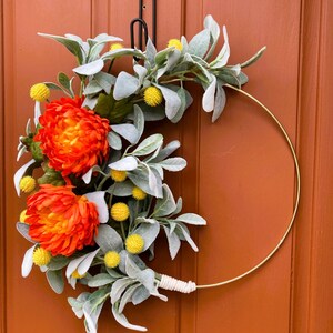 May include: A gold metal wreath with orange and yellow flowers and gray-green leaves. The wreath is hanging on a brown door.