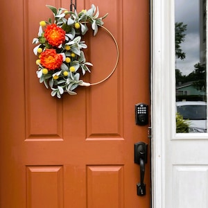May include: A wreath with orange and yellow flowers and green leaves hangs on a brown door. The door has a black door handle and a keypad lock.