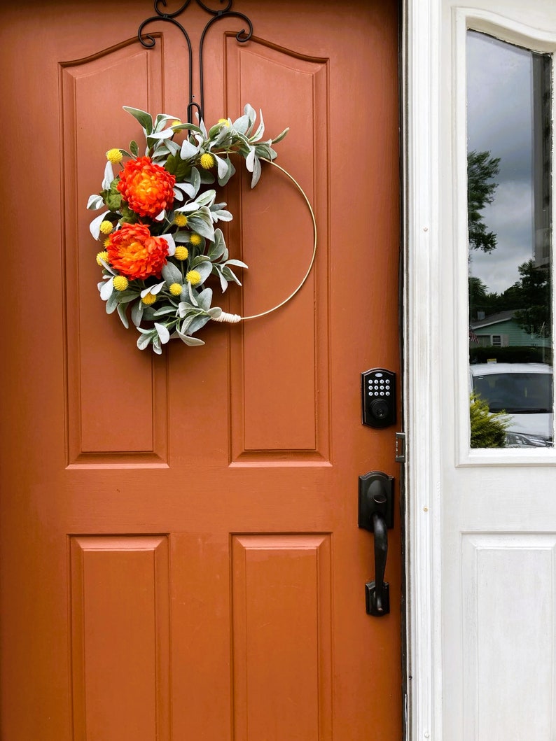 May include: A wreath with orange and yellow flowers and green leaves hangs on a brown door. The door has a black door handle and a keypad lock.
