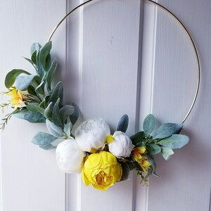 May include: A gold metal wreath with white and yellow artificial flowers. The wreath is hanging on a white door.