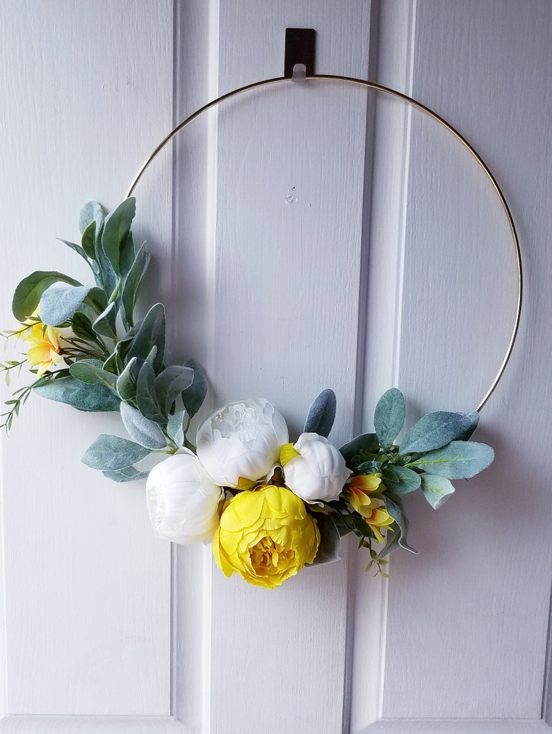 May include: A gold metal wreath with white and yellow artificial flowers. The wreath is hanging on a white door.