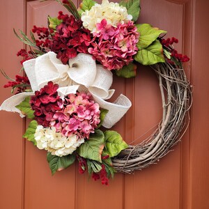 Puede incluir: Una corona de vid decorada con flores de hortensia rosa, blanca y roja, cinta de arpillera y vegetaci&oacute;n.