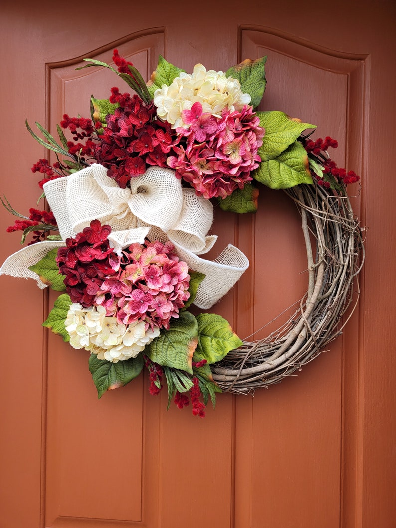Puede incluir: Una corona de vid decorada con flores de hortensia rosa, blanca y roja, cinta de arpillera y vegetaci&oacute;n.