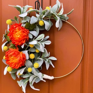 May include: A gold metal wreath with orange and yellow flowers and gray-green leaves. The wreath is hanging on a brown door.