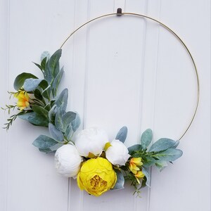 May include: A gold metal wreath with white and yellow artificial flowers. The wreath is decorated with green leaves.