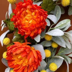 May include: A fall wreath with orange and yellow flowers and gray-green leaves. The wreath is made of artificial flowers and leaves.