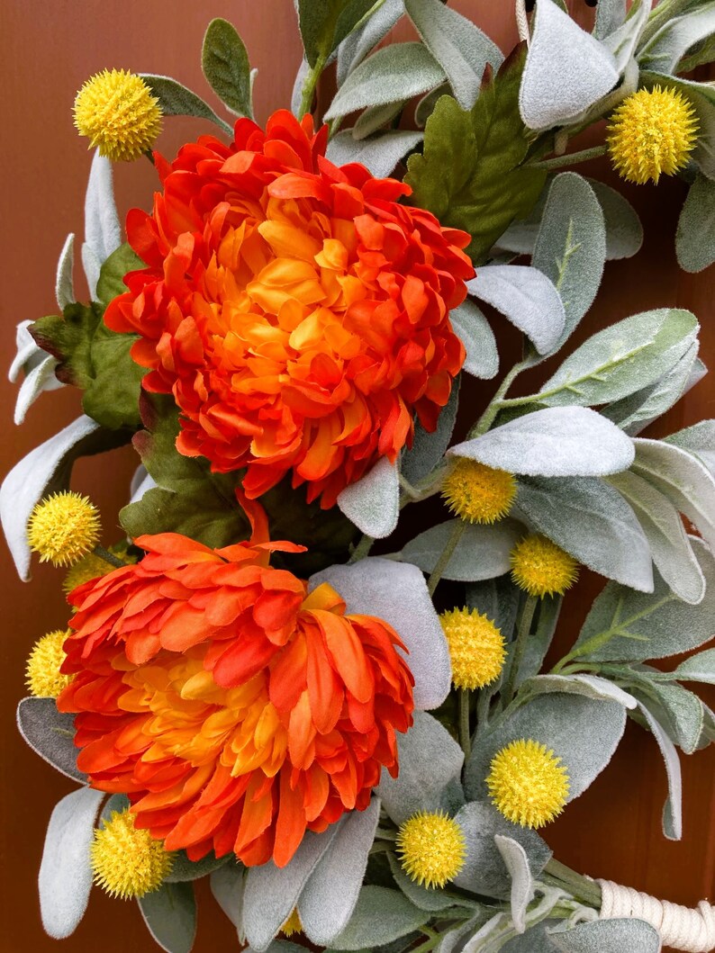 May include: A fall wreath with orange and yellow flowers and gray-green leaves. The wreath is made of artificial flowers and leaves.