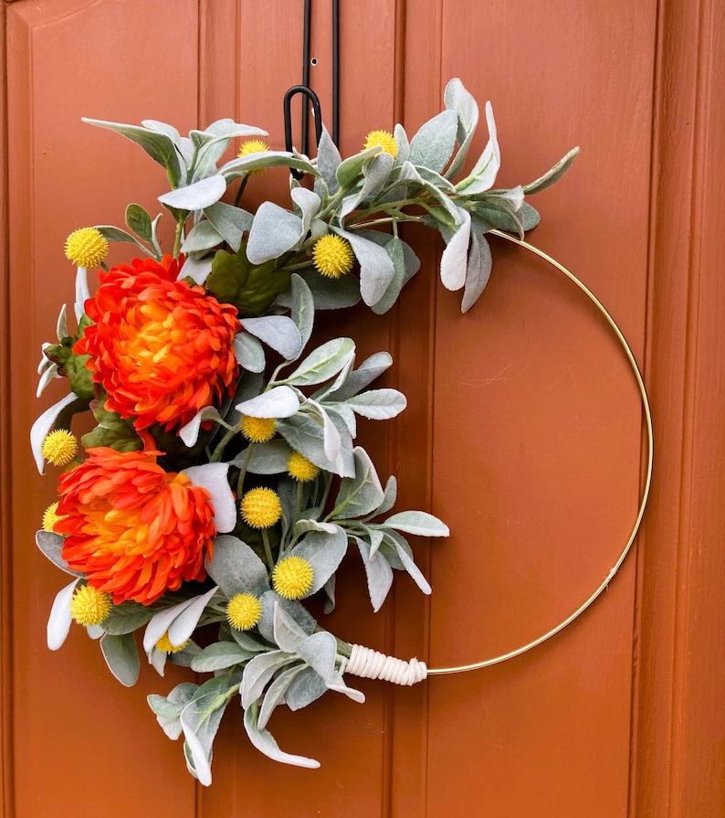 May include: A gold metal wreath with orange and yellow flowers and gray-green leaves. The wreath is hanging on a brown door.