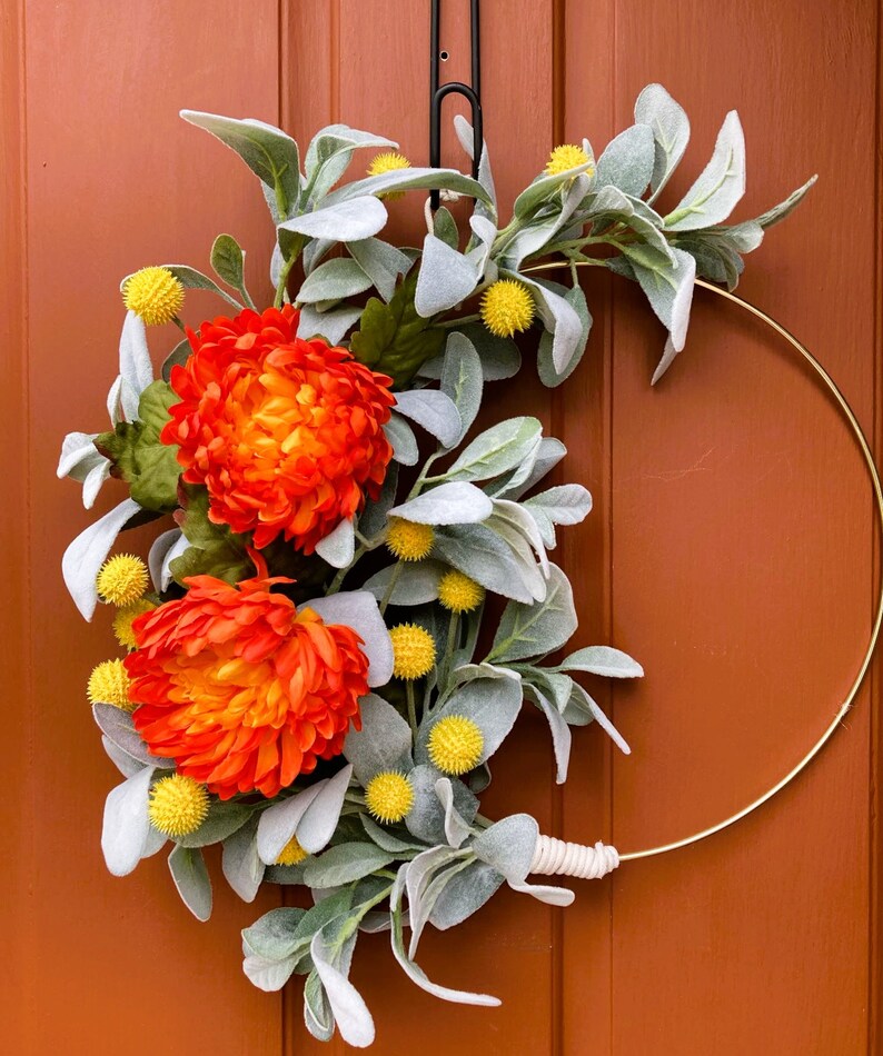 May include: A gold metal wreath with orange and yellow flowers and gray-green leaves. The wreath is hanging on a brown door.