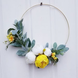 May include: A gold metal hoop wreath decorated with white and yellow artificial flowers and green leaves.
