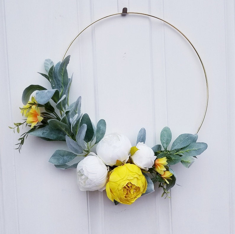 May include: A gold metal hoop wreath decorated with white and yellow artificial flowers and green leaves.